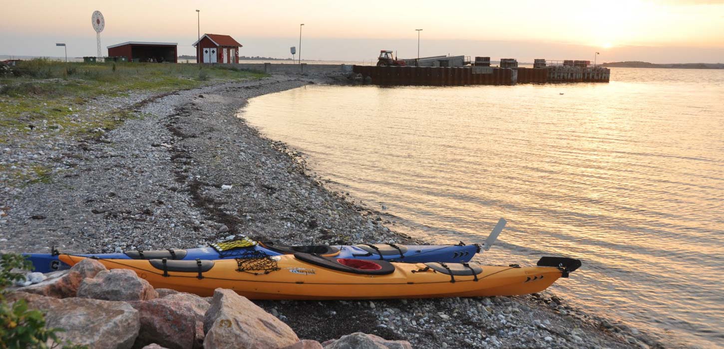 På en strandbred ligger to kajakker i forgrunden. I horisonten af billedet kan man se en strandspids hvor der står et lille rødt hus foran solnedgangen.