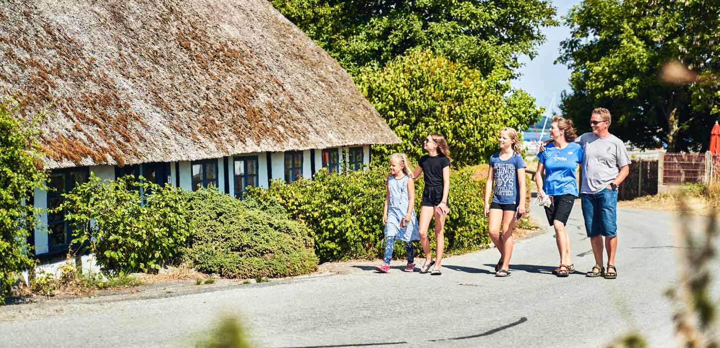 I fuld solskin går en familie med tre børn på række. De går på en asfaltvej. I baggrunden omfavner træer vejen og i venstre side ses et hvidt bindingsværks hus med stråtag. 