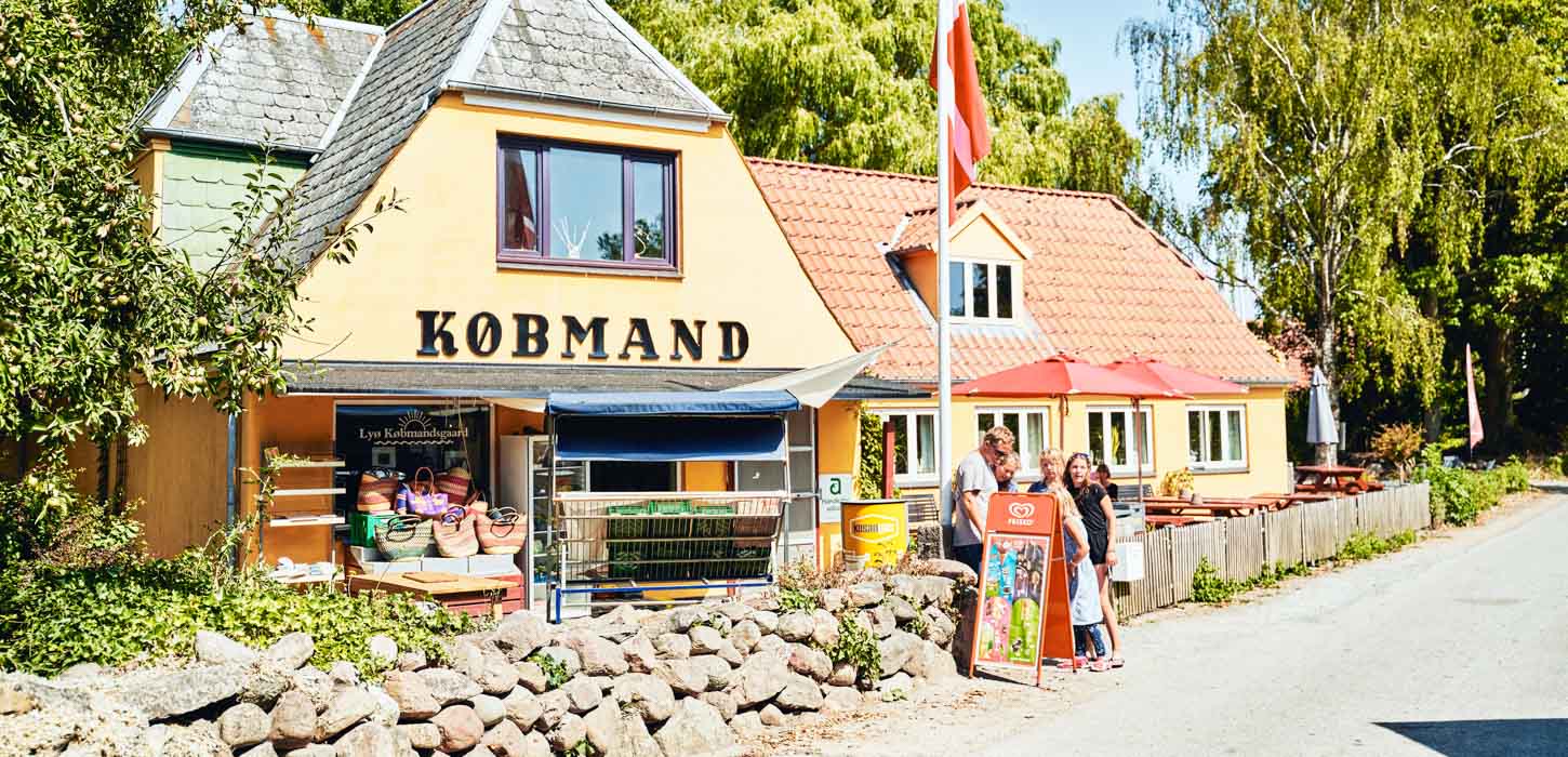 Et gult hus med rødt tag er omringet af træer. Over hoveddøren står skriften "Købmand". I forhaven står varer og en familie kigger på en is-skilt. 