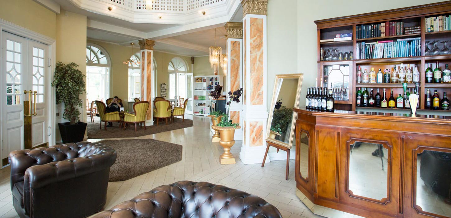 En hotellobby med lyst gulv og lyse vægge og en bar af mørkt egetræ til højre. Placeret rundt i rummet er der også mørkebrune møbler