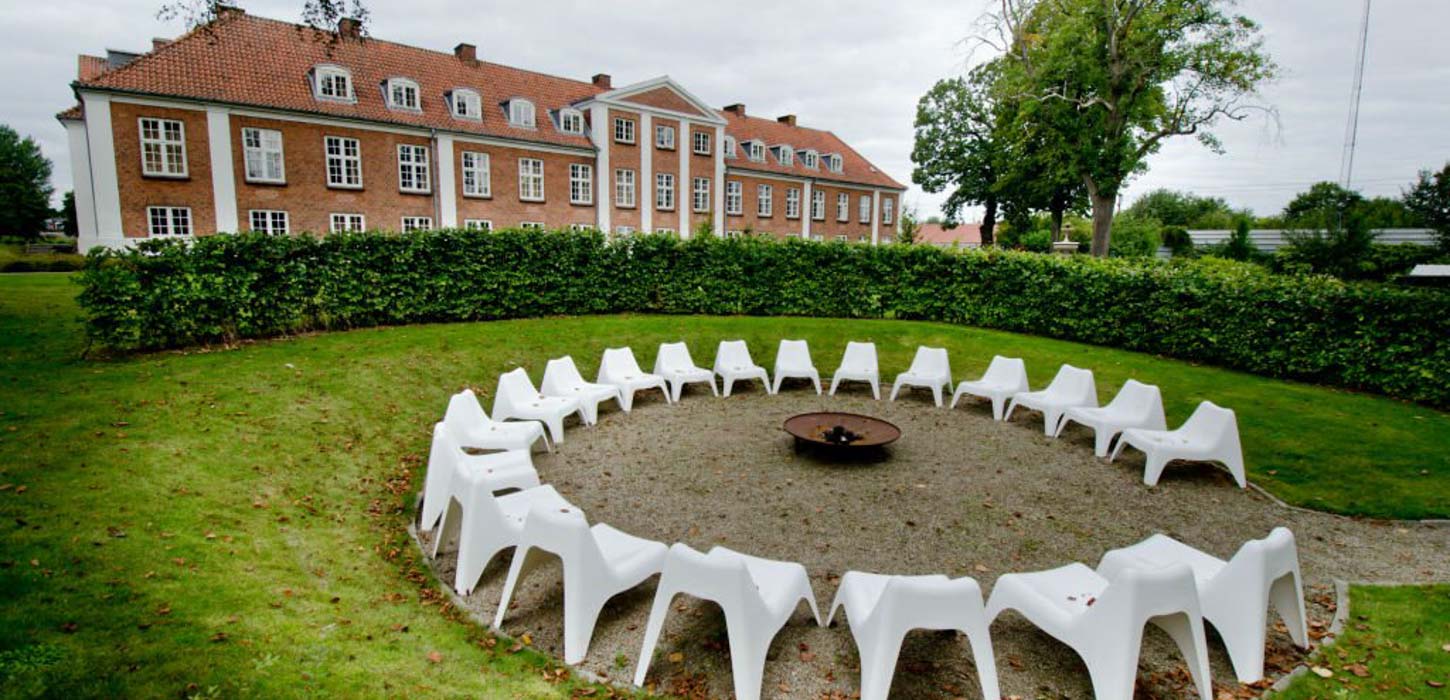 20 hvide stole stor rundt om et bålfad. Cirklen er omgivet af græsplæne og hæk. I baggrunden kan man se Hotel Parks røde mustensbygning og to træer.