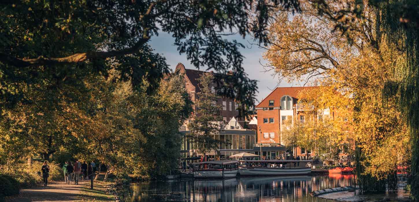 Odense Aafart på en solskinsdag i efteråret og folk går tur på stierne. Man kan se to både og vandcykler, som ligger i vandet foran Nelles bar.