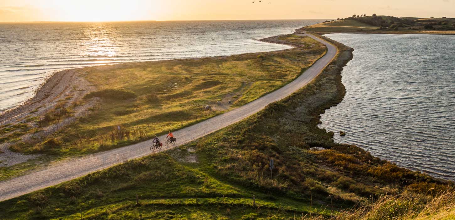 To cyklister cykler på smal grussti lige ud til vandet ved en fynsk strand. I baggrunden ses en smuk solnedgang. 