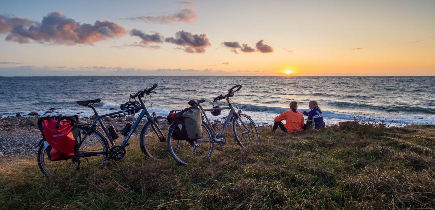 To cykler med oppakning står parkeret i strandkanten. To mennesker sidder ved siden af og ser ud over havet, hvor solen går ned.
