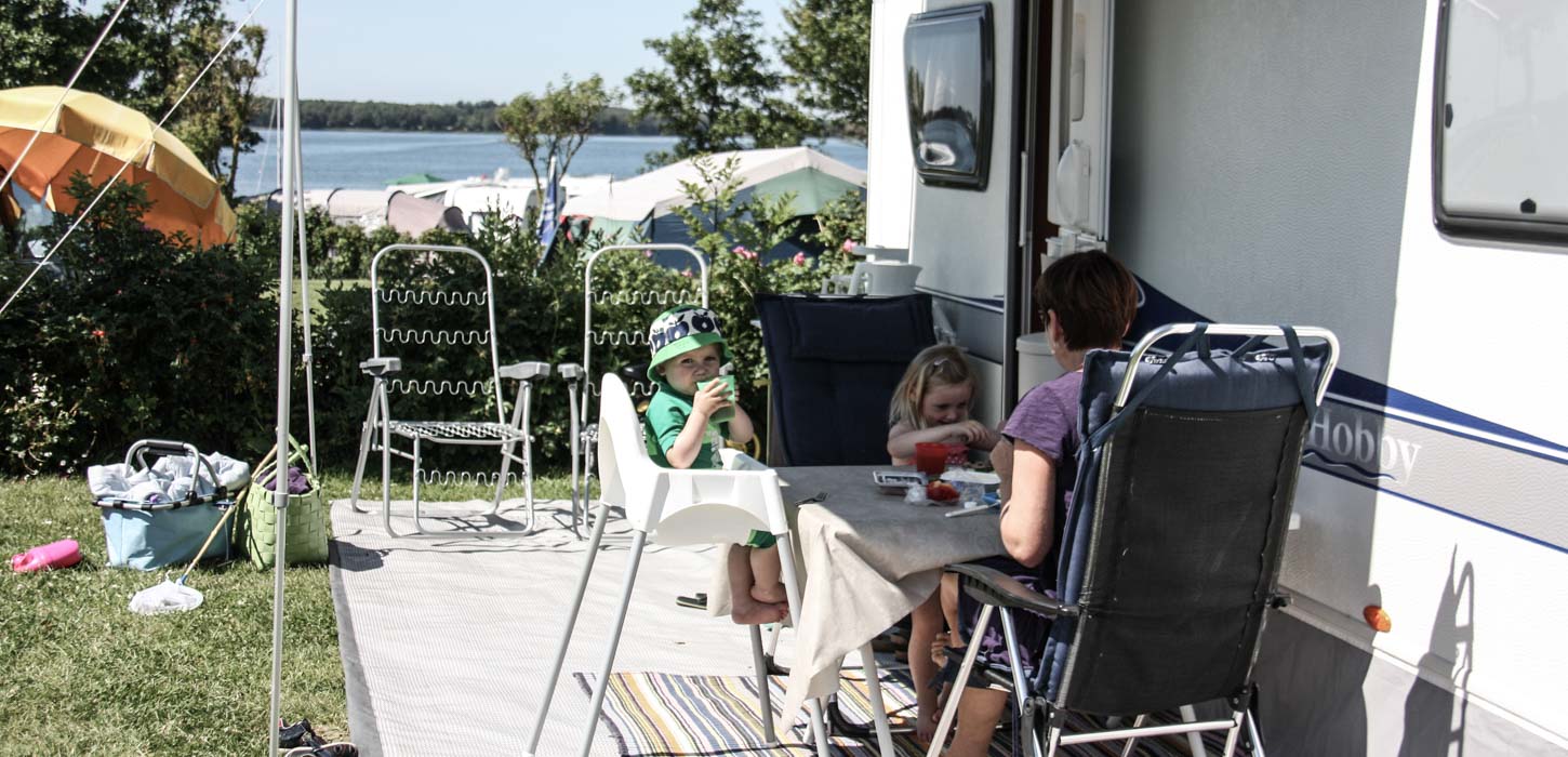 Foan campingvogn sidder en voksen og to børn i campingstole ved et bord.