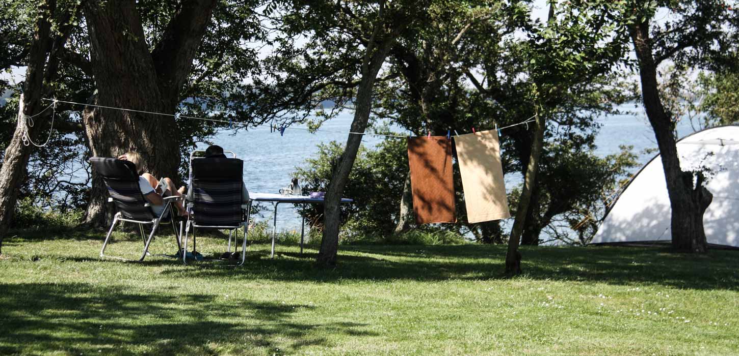 To mennesker sidder på en græsbakke på klapstol og kigger ud på havet. I baggrunden hænger to håndklæder til tørre på en snor og et hvidt telt er slået op.