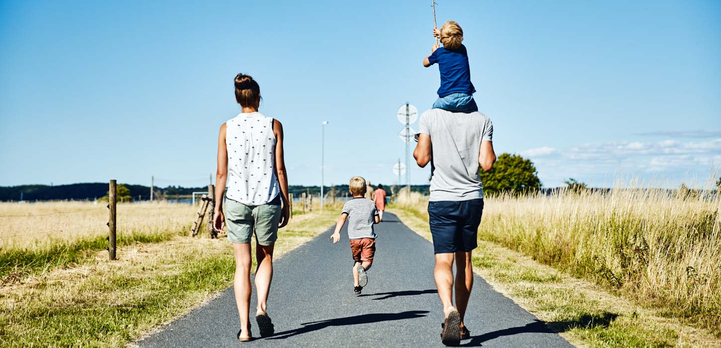 En familie på fire går ned ad en landevej på en solrig dag. Manden til højre bærer et lille barn på sine skuldre. Mellem dem går et andet barn. De er omgivet af åbne marker, og himlen er klar og blå.