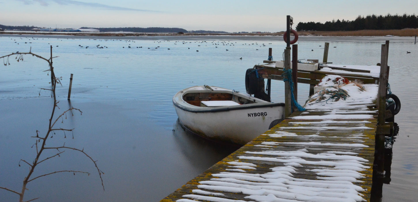 En gammel badebro har sne på sig og strækker sig ud i en stor sø og på den ene side er en jolle