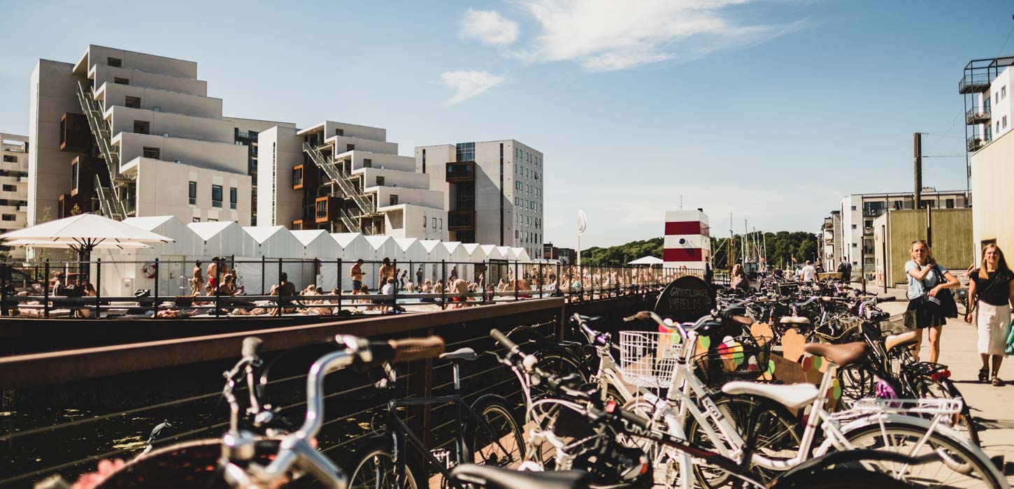 Havnebadet i Odense på en solskinsdag med blå himmel. Der er badende mennesker i havnebadet og sommerklædte mennesker, som går ude foran. Der står en masse cykler i forgrunden.