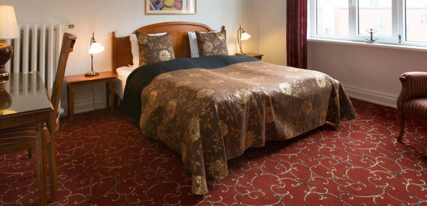 En dobbeltseng med brunt sengetæppe og sengegærde i et værelse med rødt mønstret gulvtæppe