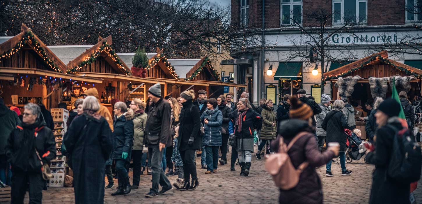 En brostensplads er fyldt med mennesker pakket ind i vintertøj. Rundt om pladsen står små træhytter pyntet med gran og julelys. Træhytterne har en åben front som giver adgang til de juleting de sælger. 