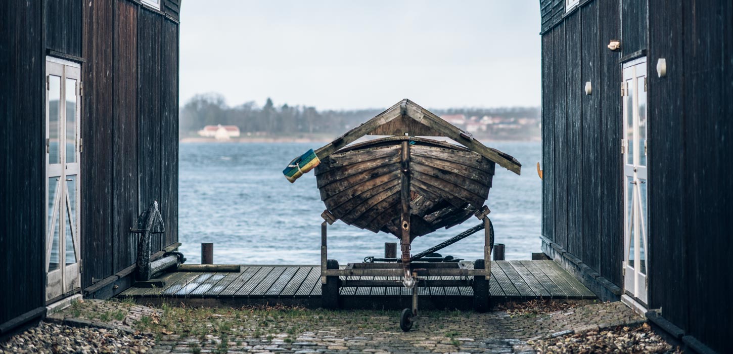 Mellem to sorte træværkshuse er en båd placeret på et stativ på havnen med udsigt til vandet. 