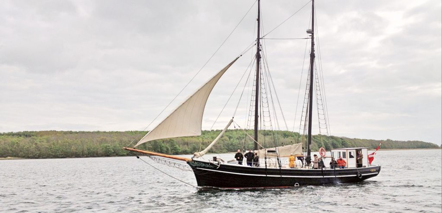 Sort sejlskib sejler over det Sydfynske Øhav med en flok mennesker ombord.