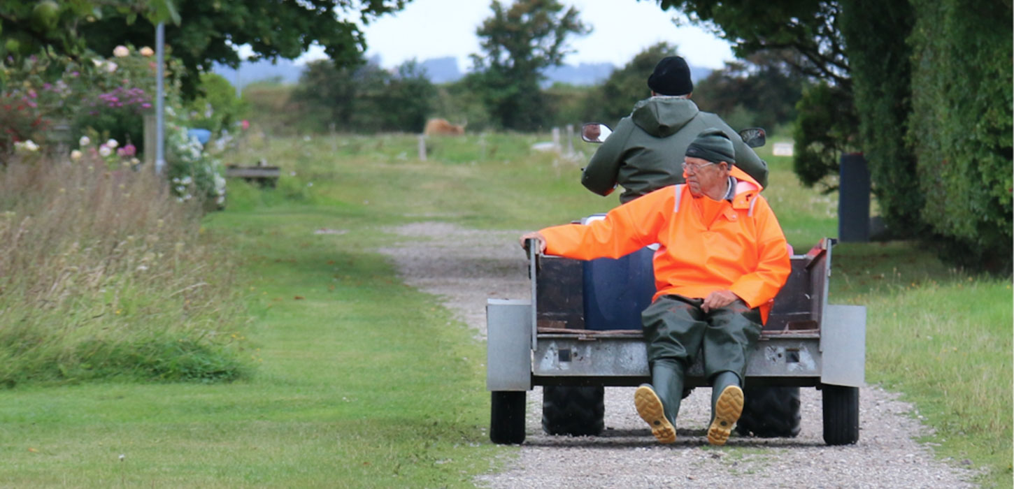 To personer på en havetraktor med trailer-lad. Manden, der kører traktoren, har ryggen til og er iklædt en grøn jakke. Den anden mand sidder bagpå på ladet med benene hængende ud over kanten i en orange jakke. De kører afsted på en grussti i grønne omgivelser.
