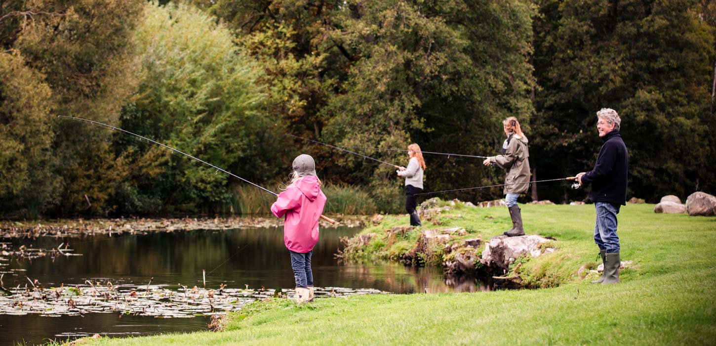 En mand, en kvinde og to børn står på en græsbeklædt flade og fisker. Der er åkander på søen og store, grønne træer i baggrunden.
