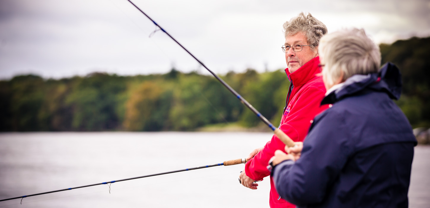 To lystfiskere står med fiskestængerne ved vandet. Den ene med ryggen til i en mørkeblå jakke, den anden kigger sig skråt over skulderen og er i en rød jakke. I baggrunden ses vandet og grøn beplantning inde på land.