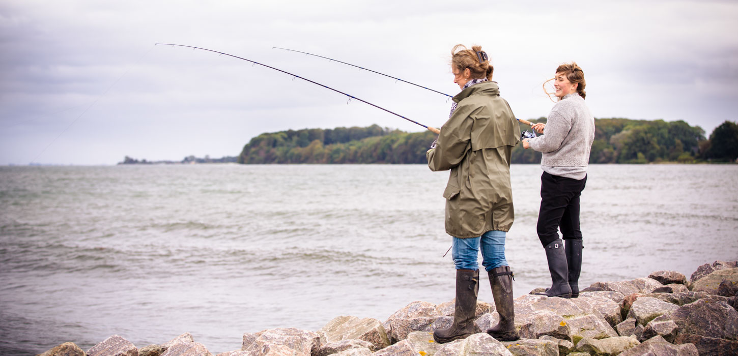 To kvinder står på store sten i vandkanten med deres fiskestang og fisker. De er iført gummistøvler , og den ene har en lang, grøn jakke på, emns den anden står i en sweater. De har begge vind i håret.