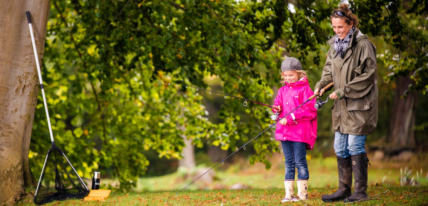 En storsmilende kvinde og en pige i pink jakke står i grønne omgivelser og fisker.