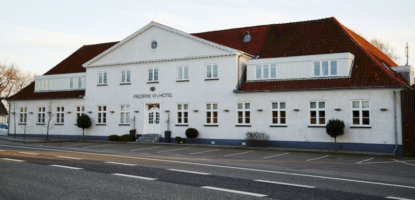 Frederik VI's hotel set ude fra vejen. Bred, hvid bygning med rødt tag ud mod asfaltvej med hvide striber.
