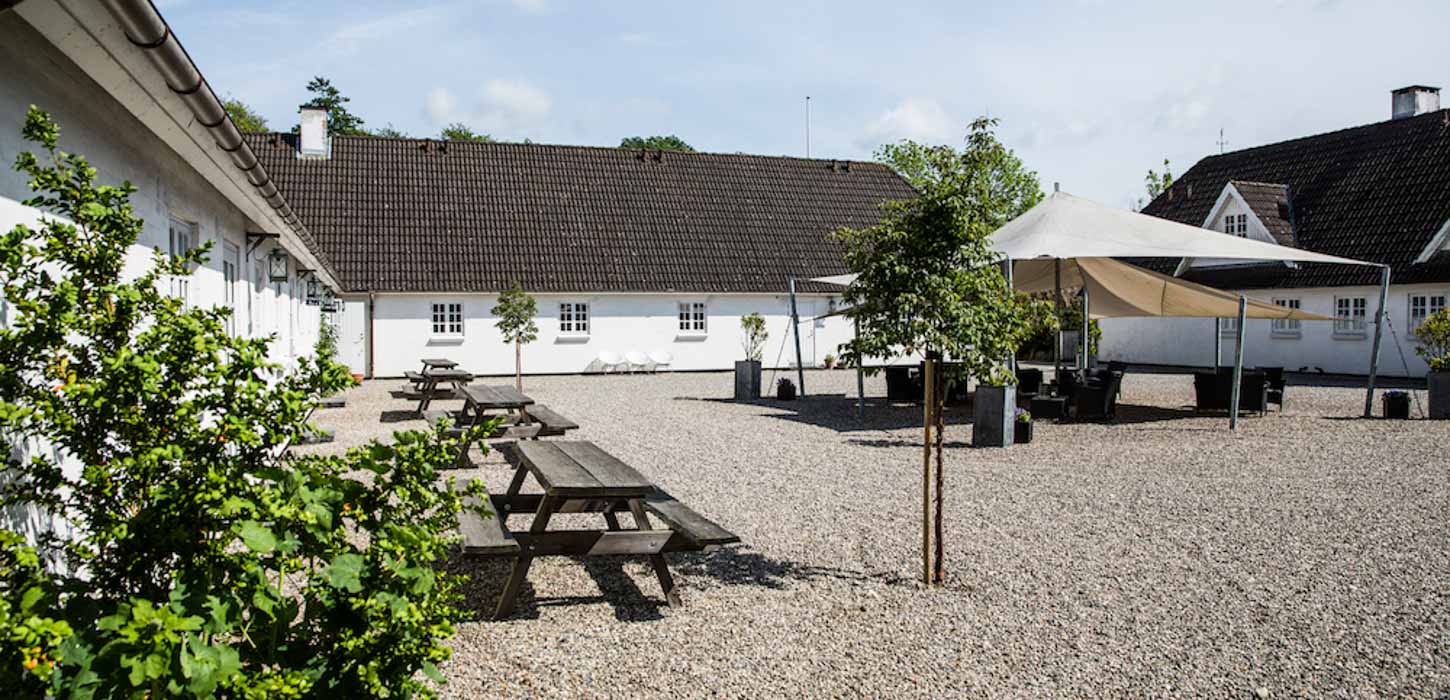 Stor gårdsplads med busk, træ, pavilloner og borde-bænke-sæt og en lang hvid bygning med tegltag i baggrunden.
