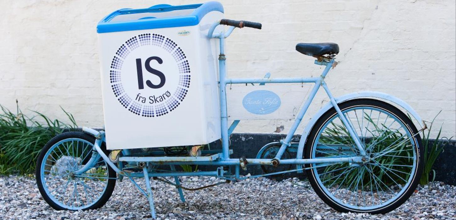 Lyseblå is-cykel med logoet "Is fra Skarø" står parkeret ved en væg.
