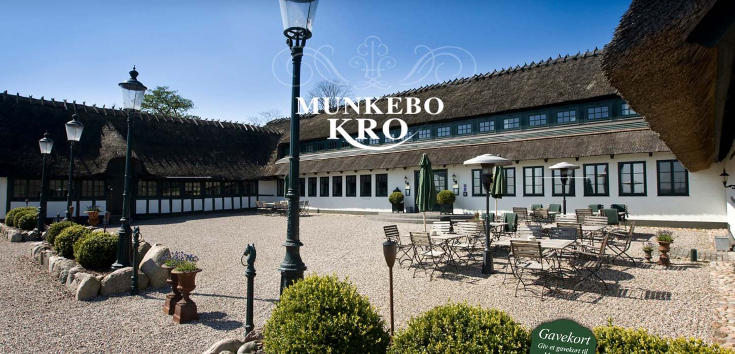 Hvid trelænget gård med stråtag, masser af vinduer og en gårdsplads med borde og stole samt små buske og lygtepæle.