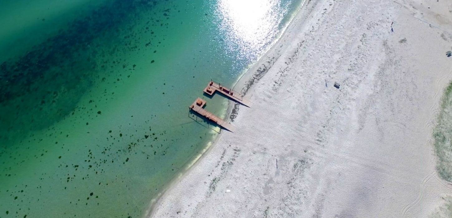 Strand set ovenfra med lyst sand og grønligt vand.