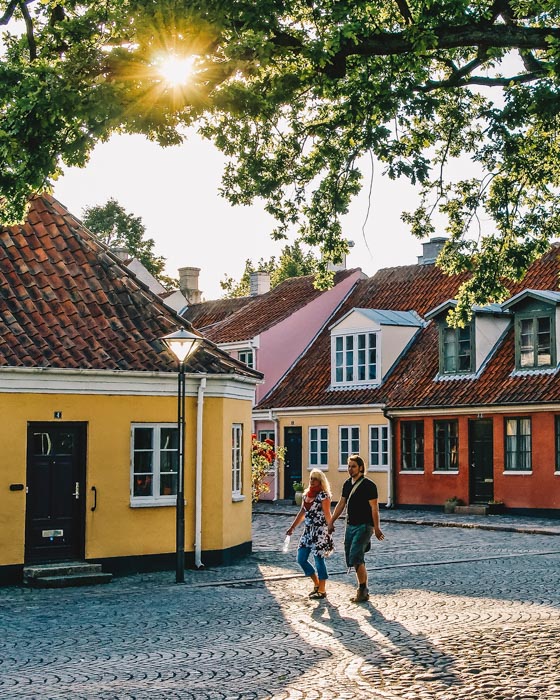 Et par går hånd i hånd på en brostensgade. I baggrundenden er fire bygninger i forskellige farver. Solen skinner gennem en trækrone. 