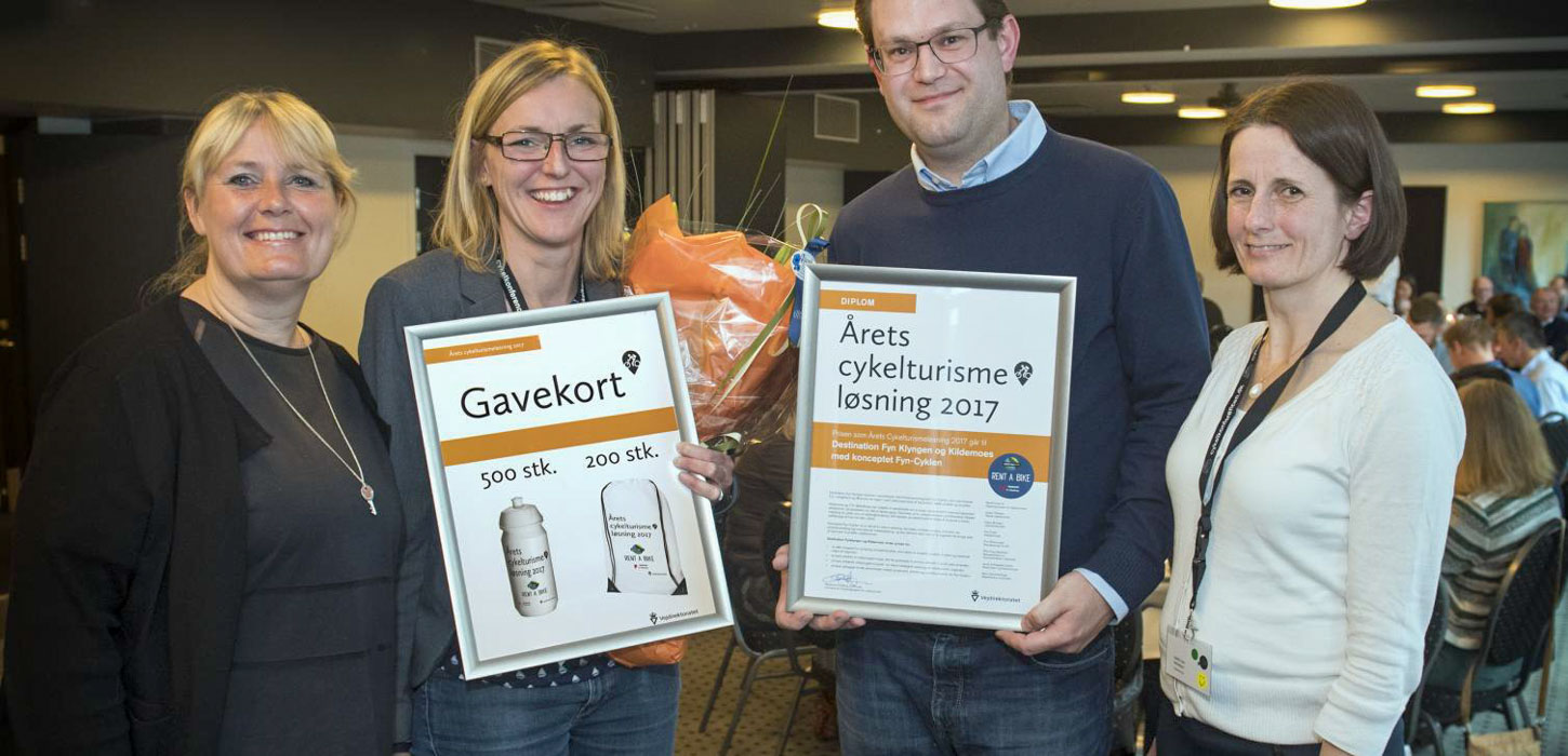 En mand og to kvinder bliver fotograferet. En mand og kvinde i midten står hver især med et indrammet billede, hvor der står 'Gavekort' og 'Årets cykelturismeløsning 2017'