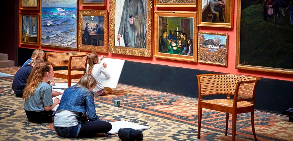 Fire personer sidder på gulvet i et kunstgalleri og tegner. De er fokuseret på deres arbejde, omgivet af en række indrammede malerier. Gulvet er dækket af et mønstret tæppe, og der er en tom stol i forgrunden.
