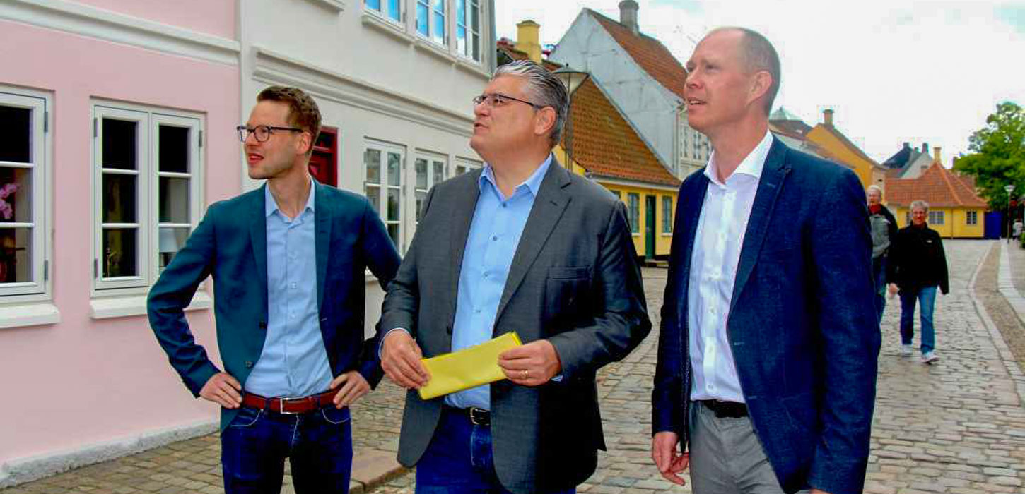 Tre mænd står side om side og ser opad, påen gade med brosten. Den første mand bærer briller og en blå skjorte, den midterste holder på et gult papir og har en grå blazer på, og den tredje bærer et mørkeblå jakkesæt. Bygningerne i baggrunden er farverige og himmelen er overskyet.