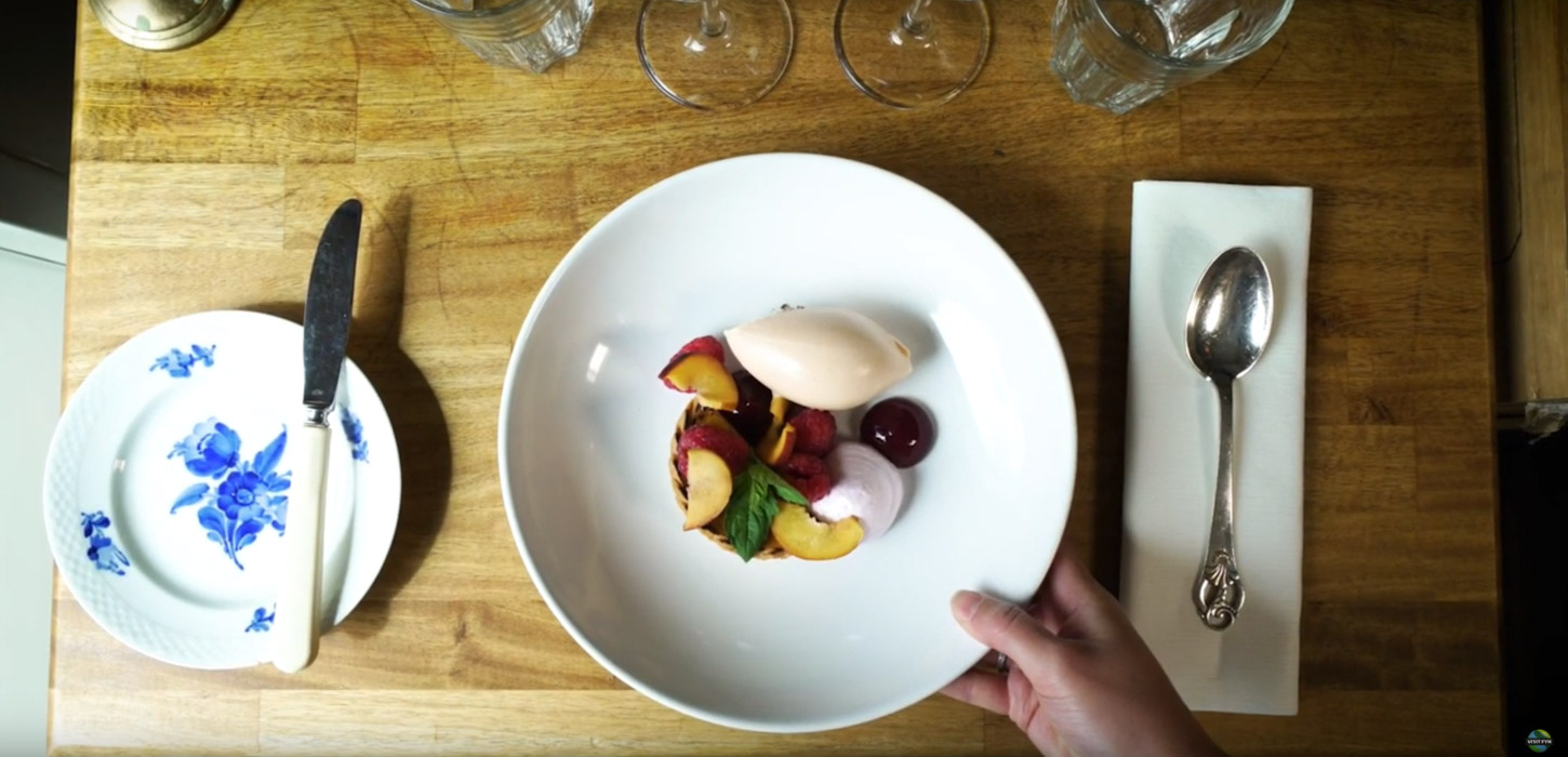 Tallerken med en velanrettet dessert på hvid tallerken og med serviet, ske, brødtallerken og kniv set oppefra. 