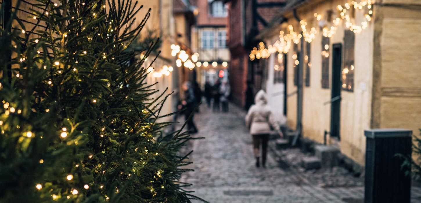 I højre side står et juletræ med julelys. Bag juletræet ses en sløret brostensgade der går ned mellem to rækker af gamle bindingsværkshuse. På husene hænger julelys, og på gaden går et par mennesker.