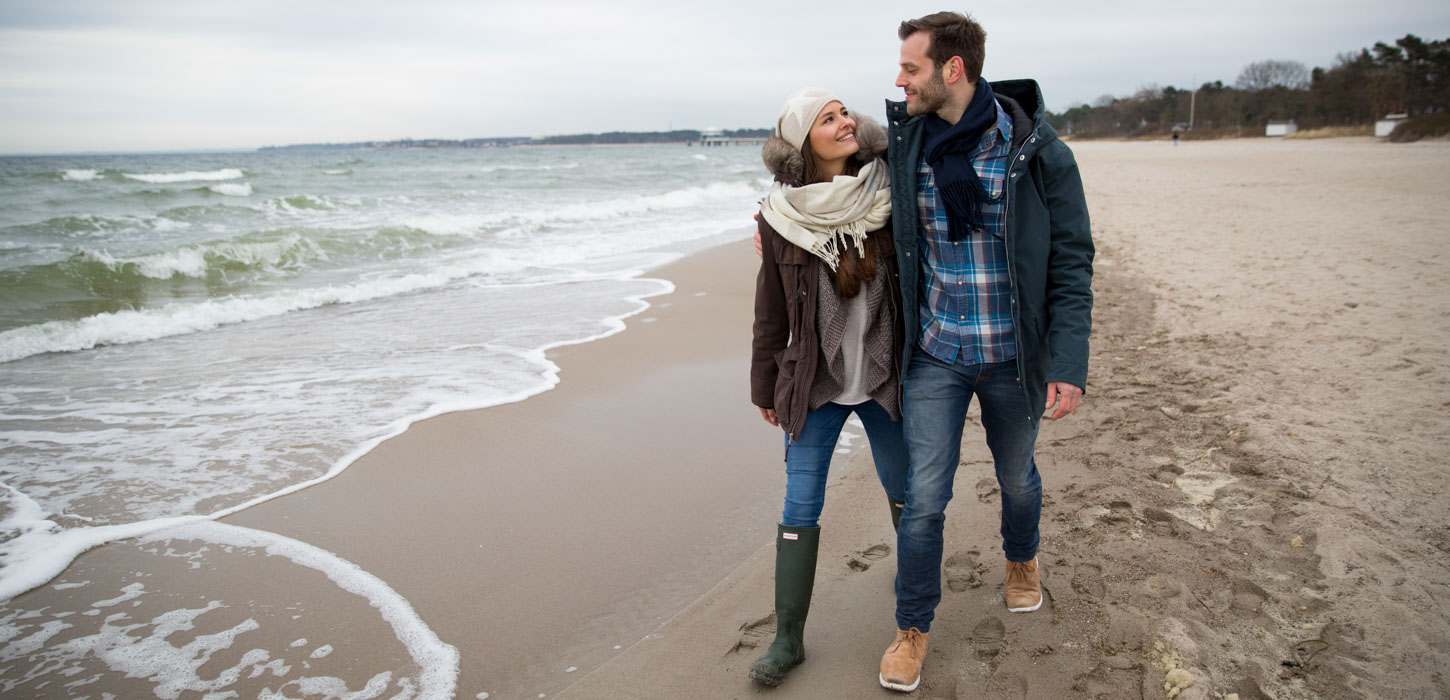 Par går langs vandkanten på stranden i tykt overtøj da det er vinter