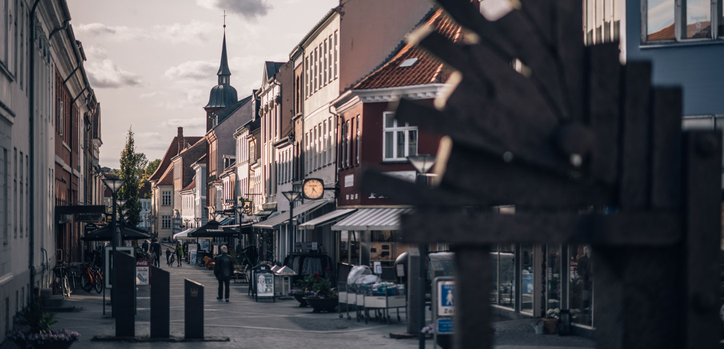 Gågade med mennesker og charmerende, gammeldags bygninger og toppen af et tårn i baggrunden.