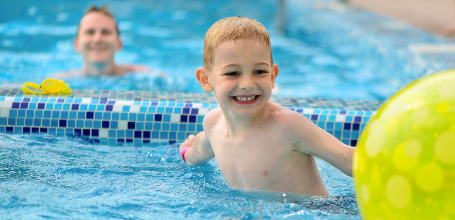 En glad dreng med et stort smil svømmer i en pool med blå mosaik-fliser. Bag ham anes en voksen, der smilende ser på barnet. En del af en stor grøn badebold er synlig i forgrunden.
