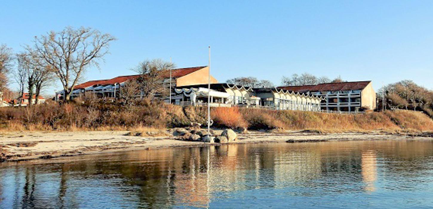 En stor rød bygning med glaspartier ligger ud til vandet. Vandet er stille og solen skinner.