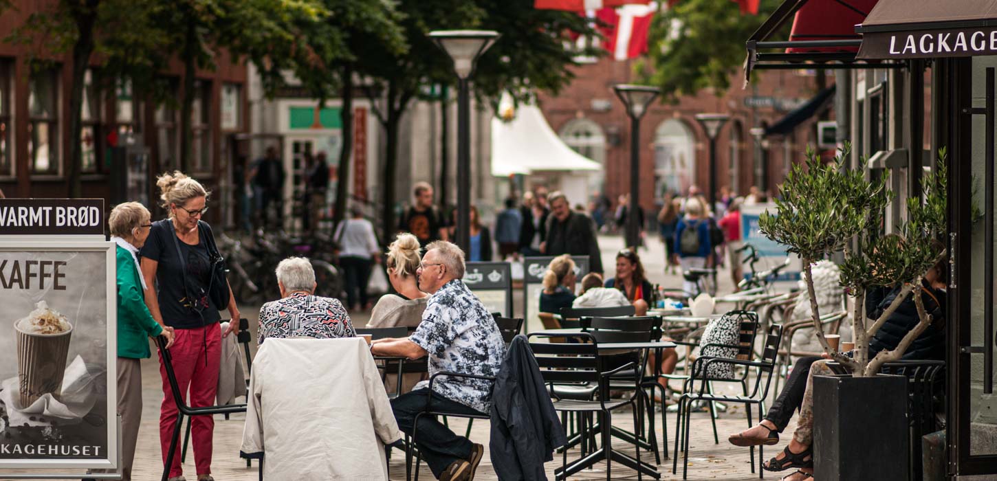 Mennesker sidder udenfor foran Lagkagehuset i Odense gågade. I venstre side står der er et skilt med med et billede af en kop varm kaffe. 