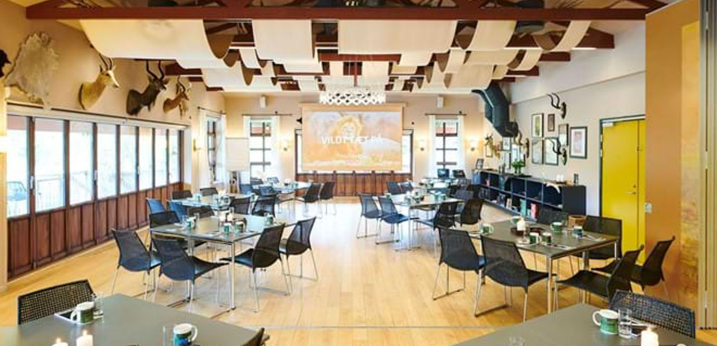 Mødelokale i brune, gule og beige farver med borde, stole og skærm. Bordene er inddelt i grupper med plads til seks mennesker ved hvert. Rundt omkring hænger der udstoppede dyrehoveder på væggene.