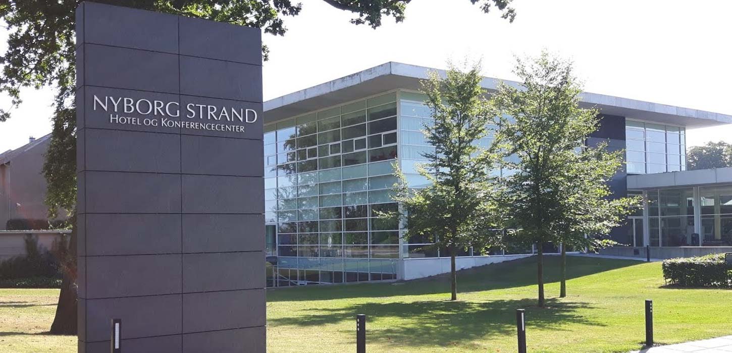 På et højt skilt står der Nyborg Strand Hotel og Konference. Bagved skiltet kan man se en græsplæne med tre træer der står foran en hvid firkantet bygning med mange vinduer.