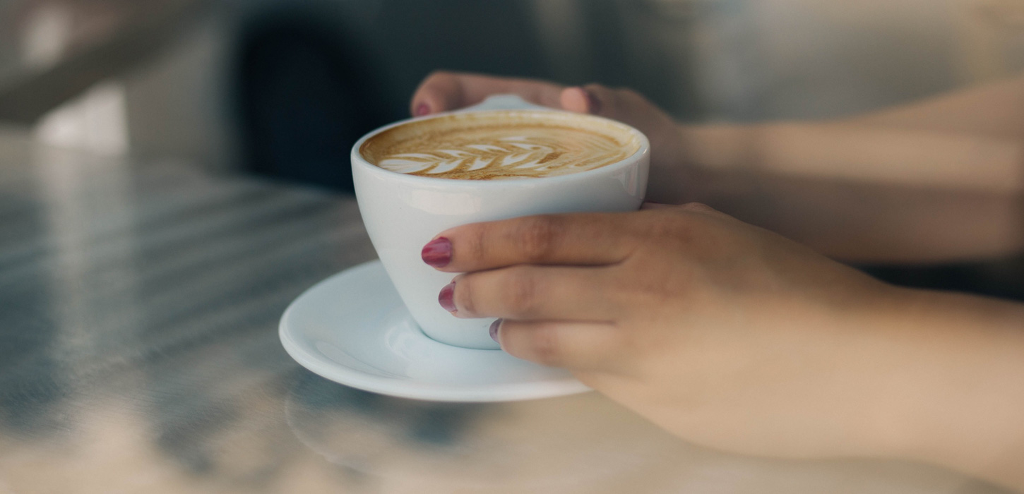 Gennem et vindue er der taget et nærbillede af to hænder der holder en kop kaffe. Hænderne har lyserød neglelak på. Hænderne holder om en hvid kop. I koppen er der kaffe og den er pyntet med latte-art der ligner et blad. Koppen står på en hvid underkop, som står på et lyst træbord.