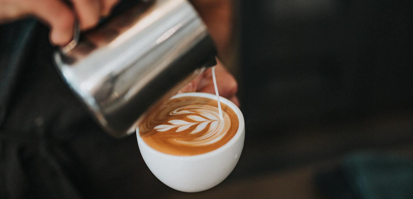 To slørende hænder er ved at lave en kop barista kaffe. I den ene hånd er en hvid kaffe kop fyldt til kanten med kaffe. I den anden hånd er en sølvkande. Fra kanden hældes der mælk ned i koppen som former et blad.