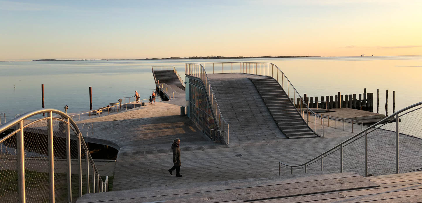 En stor badebro med udspringsmuligheder og havet i baggrunden, der genspejler en blå og gullig himmel