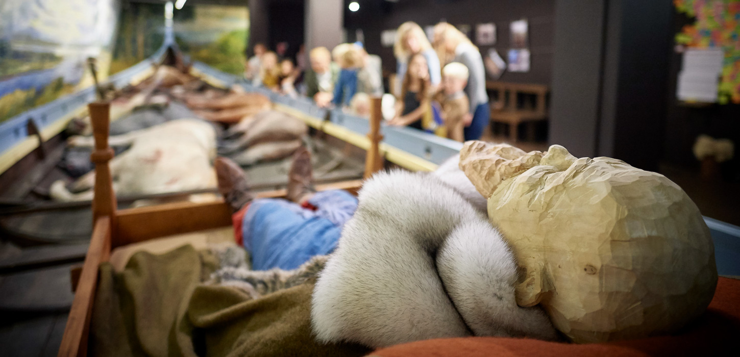 Et billede taget fra den ene ende af et vikingeskib. I forgrunden ligger en træfigur i en træseng iklædt vikingetøj. Længere nede i båden ligger falske ulve. I højre sidde står en gruppe mennesker og kigger på udstillingen.