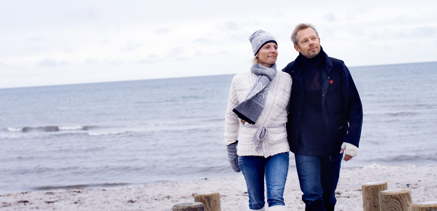 Et par går og holder om hinanden i deres vintertøj mens de kigger fremad og bagved dem er stranden og havet