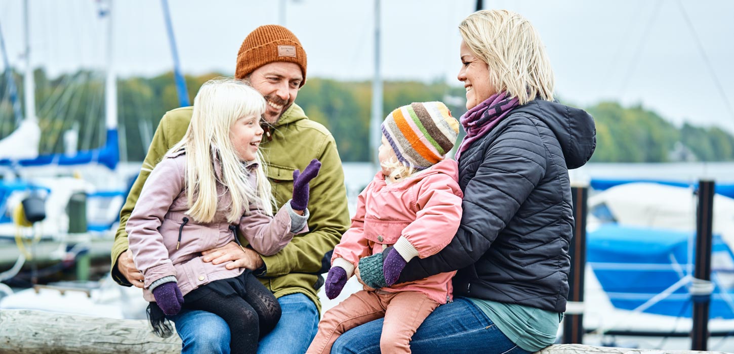 En mand og en dame sidder med hvert sit barn på skødet og i baggrunden er en havn