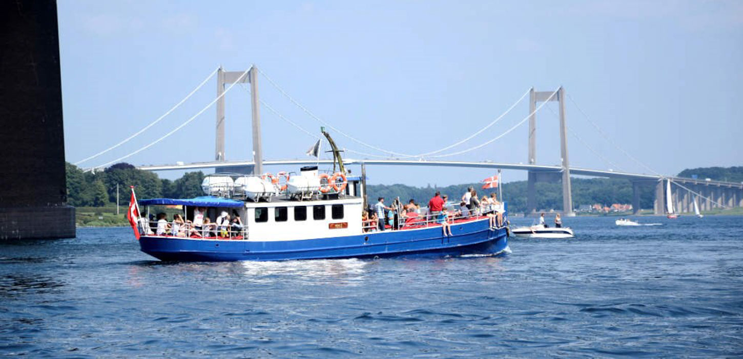 På havet foran Lillebæltsbroen sejler et blåt træskib med hvidt førerhus. På dækket står en flok mennesker og kigger udover vandet, og i bagenden flagre det danske flag.