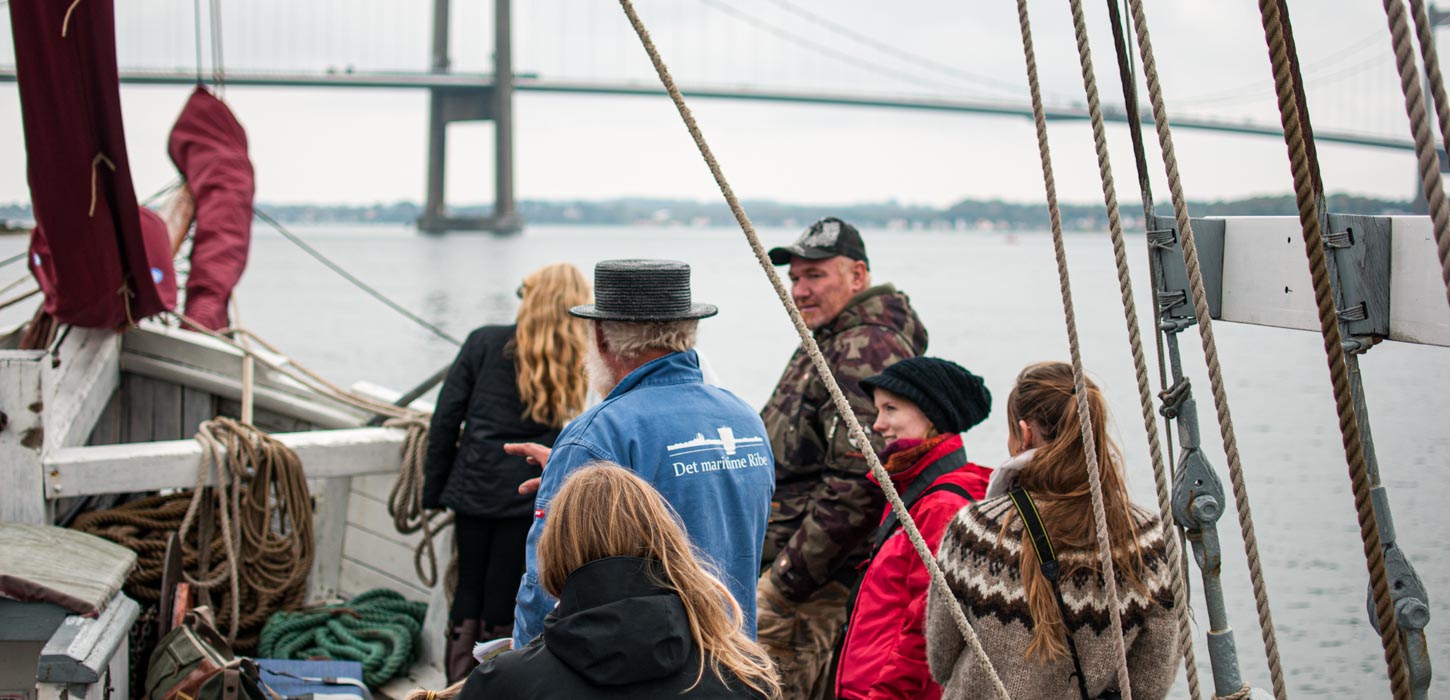 Et billede taget mellem rebbene på et sejlskib. 5 turister lytter med mens en guide med høj hat snakker. I baggrunden ses havet og Lillebæltsbroen.