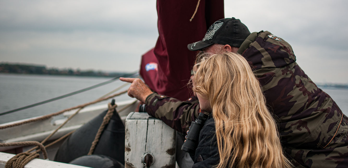 På en båd står en lyshåret pige og en mand i med kasket og camouflage jakke og kigger ud over vandet. Manden peger i den retning de kigger.