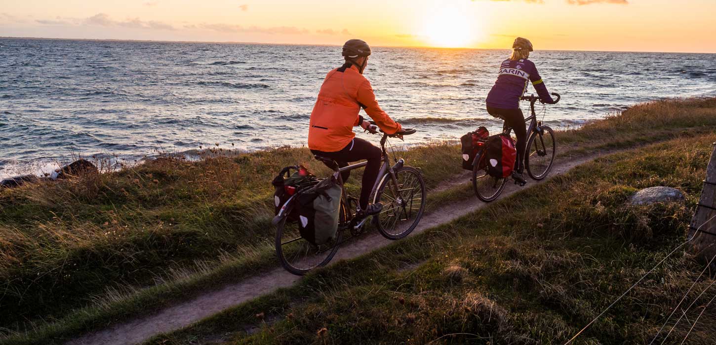 To cyklister cykler på smal grussti lige ud til vandet ved en fynsk strand. I baggrunden ses en smuk solnedgang. 